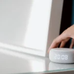 Écran connecté Apple en 2026 : Siri et Gemini, l’addition floue Close-up of a hand adjusting a smart speaker displaying time indoors.