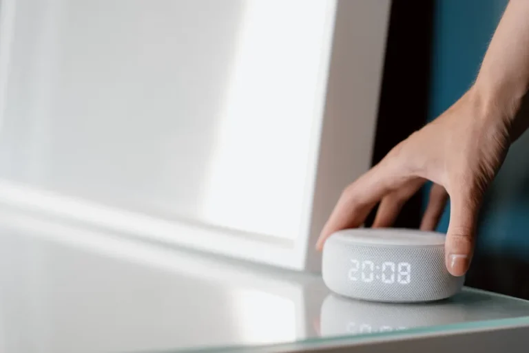Écran connecté Apple en 2026 : Siri et Gemini, l’addition floue Close-up of a hand adjusting a smart speaker displaying time indoors.