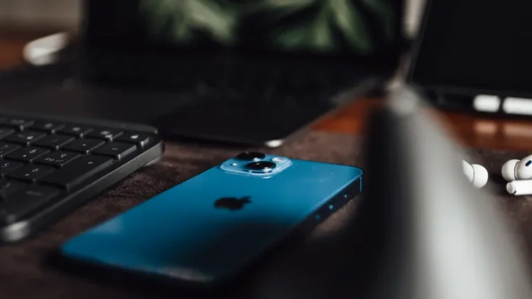 Close-up of an Apple iPhone, keyboard, laptop, and AirPods on a desk.