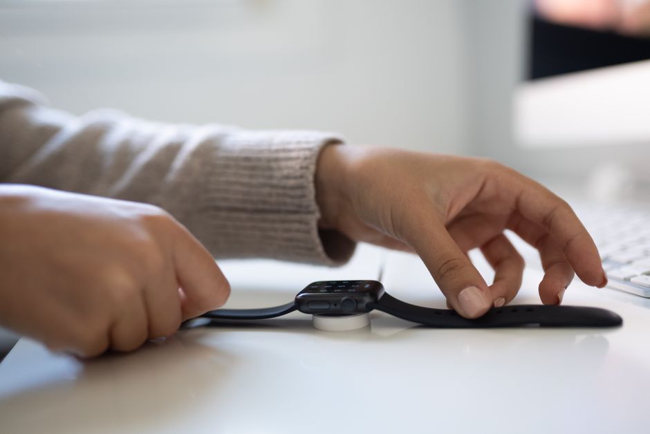 Close-up of hands placing a smartwatch on a charging dock on a white desk indoors.