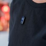 Apple préparerait un pin AI en 2027 : 20 millions visés Close-up view of a wearable tech device clipped on a black t-shirt against a blurry background.