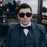 Confident man in sunglasses and bowtie standing on a city rooftop, showcasing elegance and sophistication.