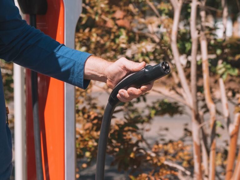Bluetti au CES 2026 : 1 200 W depuis la voiture, avec un hic Hand holding an electric vehicle charger at a station outdoors with autumn trees.