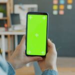 Grok bloque les faux nus le 15 janvier 2026, Apple sous pression Hands holding a smartphone with green screen in a modern office setting.