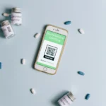iPhone 18 Pro attendu en septembre 2026 : la Dynamic Island rétrécirait Image of a smartphone displaying a health passport alongside COVID-19 vaccine vials and pills on a light blue background.
