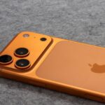 RAM plus chère : Apple compte sur l’iPhone 17 avant le 29 janvier Orange smartphone with dual cameras placed on a soft gray fabric surface.