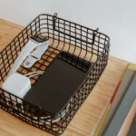 CES 2026 : Pepcom coupe une batterie de 10 000 mAh en deux Organized workspace with tech gadgets in a wire basket on a wooden desk.