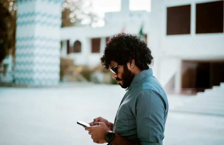 Apple en 2026 : Siri, Gemini et l’iPhone 18 jouent gros Side view of a curly-haired man with sunglasses using a smartphone in a sunny outdoor setting.