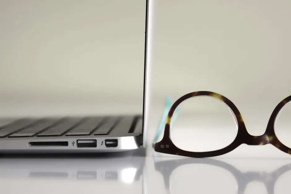 Side view of a modern laptop alongside stylish tortoiseshell glasses on a reflective surface.