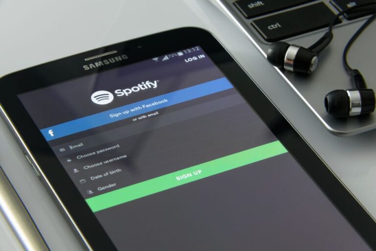 Spotify Premium passe à 12,99 $ en février 2026, Apple Music coûte 2 $ de moins Smartphone showcasing Spotify login screen beside laptop and earphones on desk.