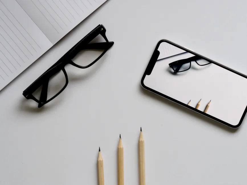 Stylish minimalist flat lay with smartphone reflection of glasses and pencils on white background.