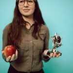 Woman holding an award trophy and apple against a turquoise background, symbolizing achievement and balance.
