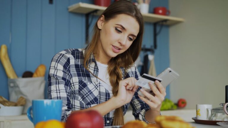 Woman in plaid shirt using credit card with smartphone in cozy kitchen setting.
